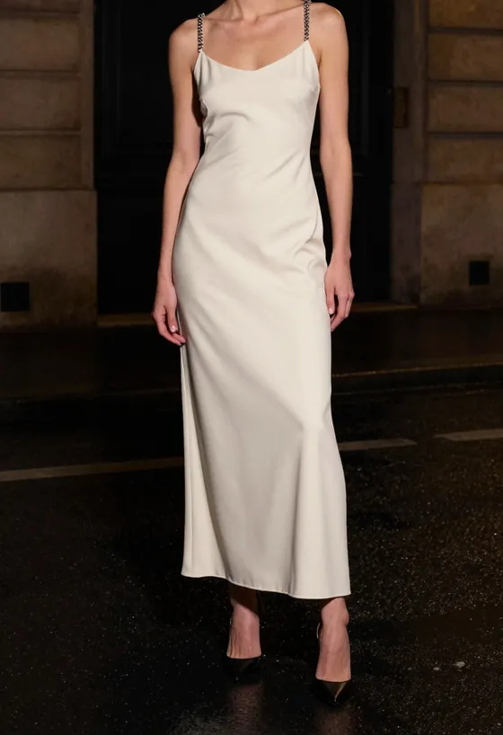 A model facing the camera, wearing a floor-length bone ivory bias-cut slip gown with thin chrome chain straps, on a wet Paris street at night.