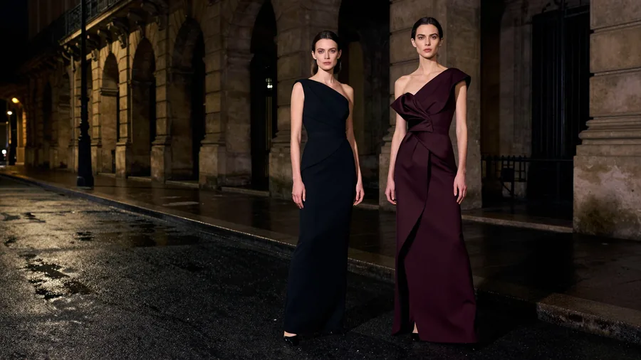 Two models in sculptural black and oxblood evening dresses under a dark stone arcade at night.