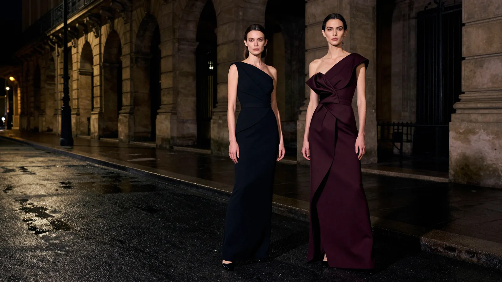 Two models in sculptural black and oxblood evening dresses under a dark stone arcade at night.