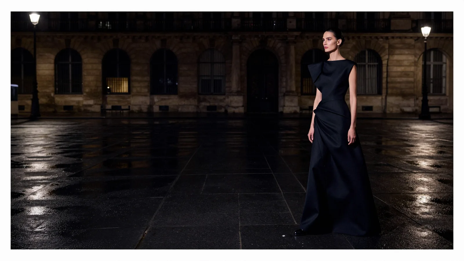 A model in a long black sculptural gown seen at a distance across an empty wet plaza at night.
