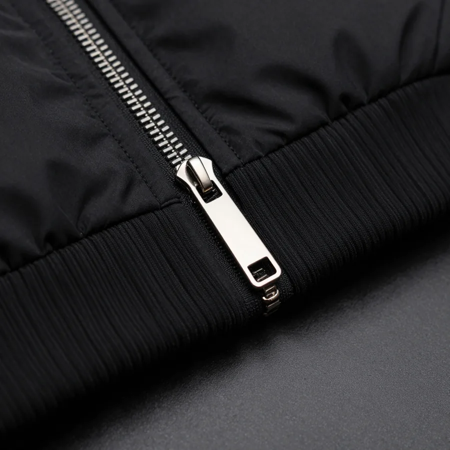 Close macro detail of the material and construction of a cropped matte black bomber jacket with polished P-formed hardware.