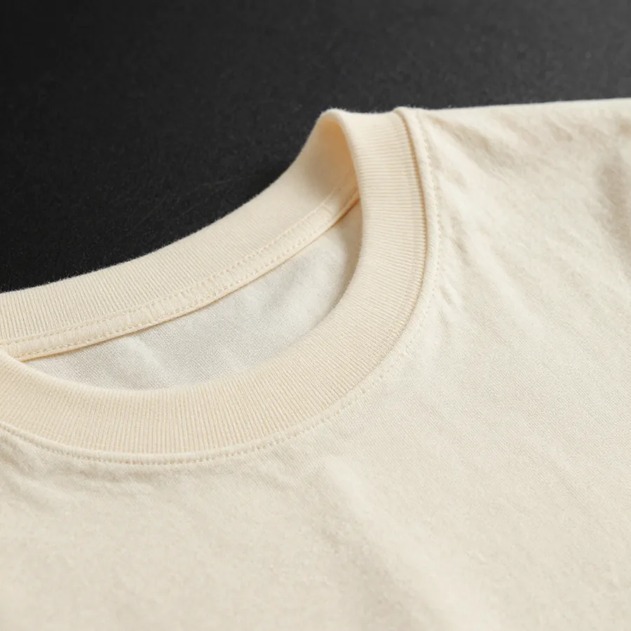 Close macro detail of the material and construction of a heavyweight bone ivory cotton t-shirt with a small gold P emblem at the chest.