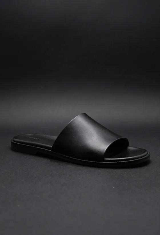 Studio product shot, side profile, of a black leather slide sandal with the P embossed into its single wide strap, on a dark ground.