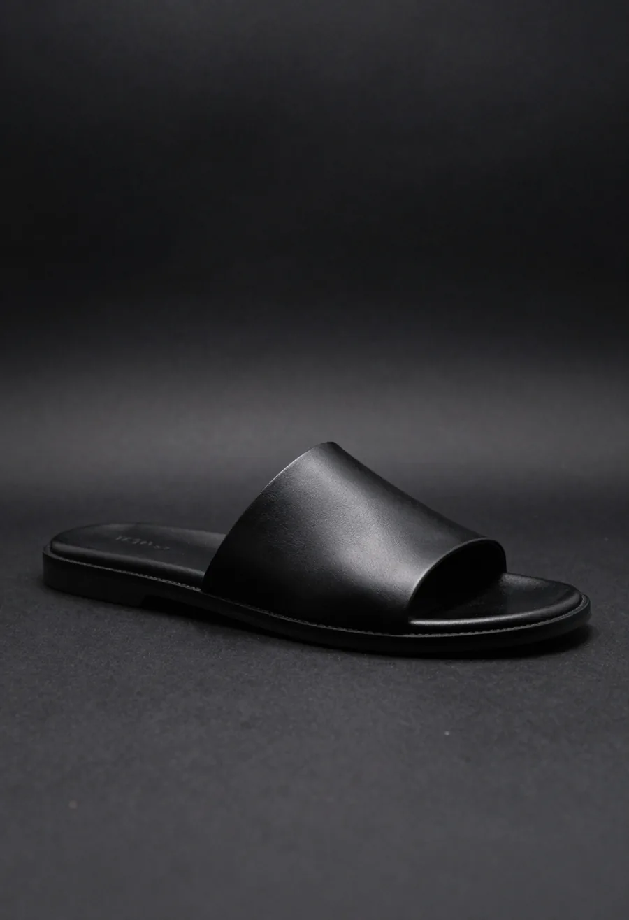Studio product shot, side profile, of a black leather slide sandal with the P embossed into its single wide strap, on a dark ground.