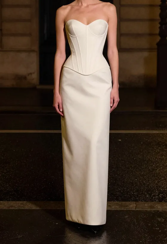 A model facing the camera, wearing a bone ivory strapless corset gown with a continuous column skirt, on a wet Paris street at night.