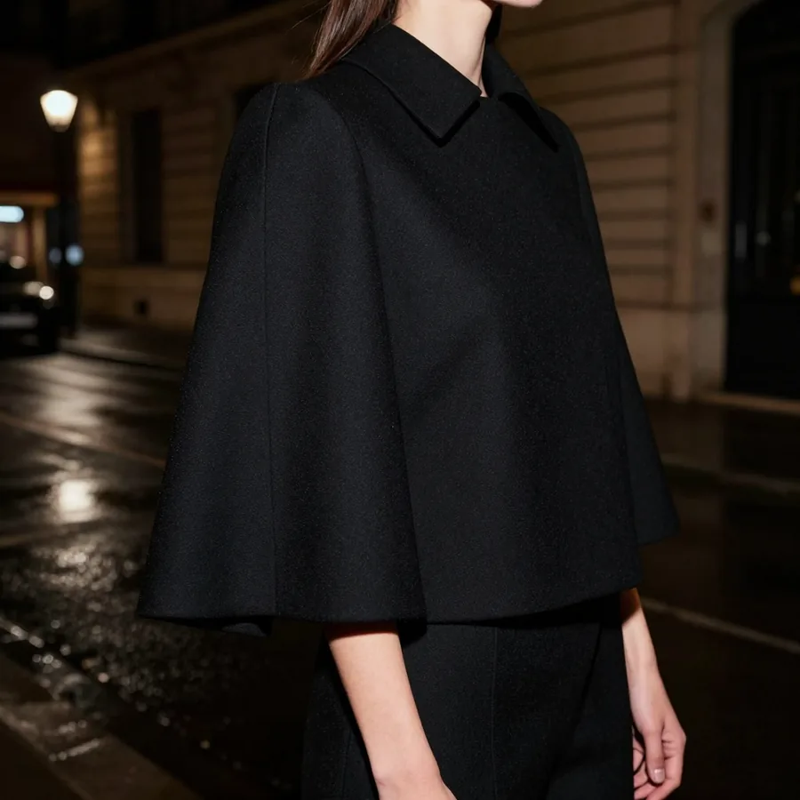 Close detail crop of the construction of a black structured cape-jacket hybrid with a sculptural shoulder, no face visible.