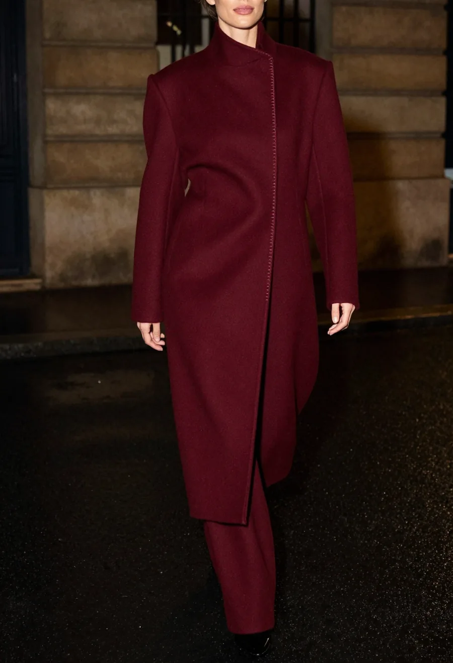 A model wearing a floor-brushing oxblood wool overcoat with the trademark house seam running the full front panel on a wet Paris street at night.